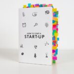 A book titled "How to Start a Start-Up" with numerous colorful sticky notes marking various pages, showcasing its extensive content. One of the best startup books for entrepreneurs looking to gain insights into launching a business.